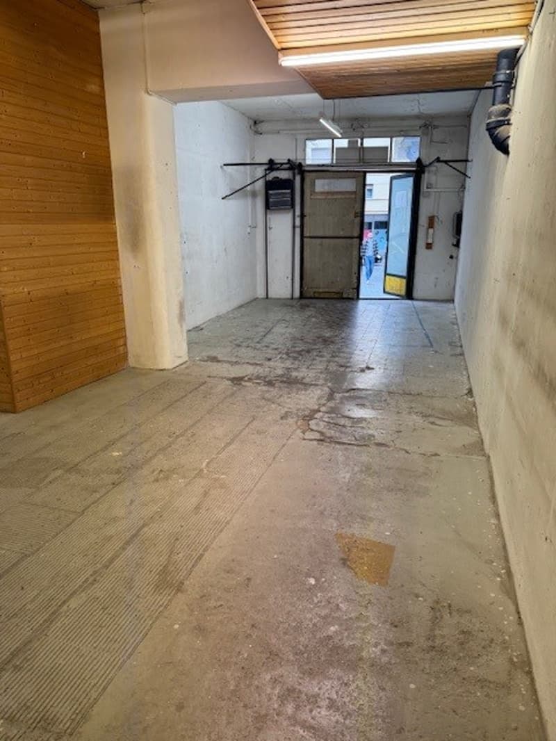The image shows a long, narrow hallway with concrete flooring and wooden paneled walls. There are various storage units and shelves lining the walls, and the hallway appears to lead to a doorway at the end.