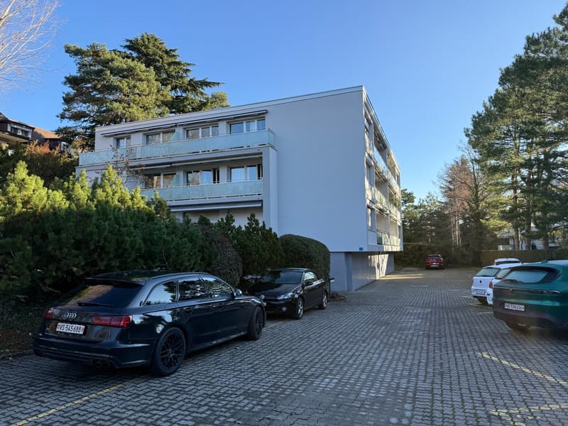 Modern apartment building, multiple floors, white facade, glass windows, balconies, surrounded by trees and bushes, paved parking area
