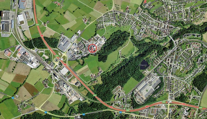 Aerial view of a town with roads, buildings, and green areas. A red circle highlights a specific building.