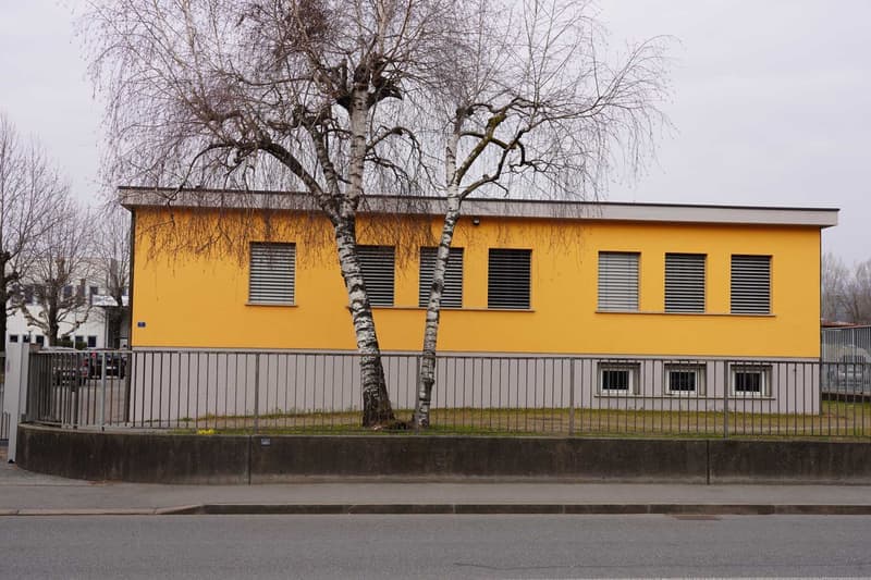 one story building, yellow facade, metal fence, two windows on first floor, large windows with shutters on second floor, two trees