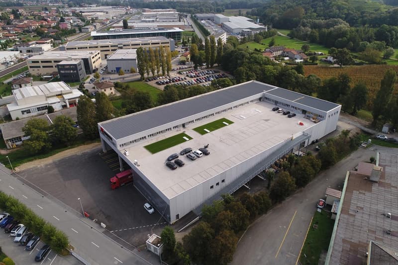 Large industrial building with parking area, roof solar panels, surrounded by other buildings and trees.