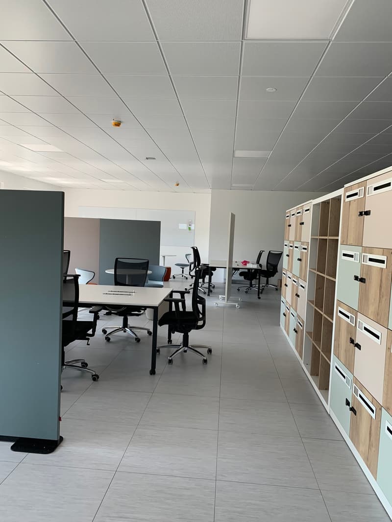 The office space has white ceiling, white walls, tiled floor, cubicles, desks, chairs, whiteboards, and storage shelves.