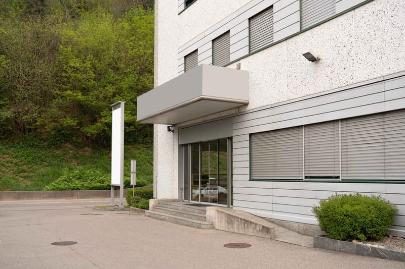White building with glass doors and windows, steps leading to entrance, security camera, greenery in the background