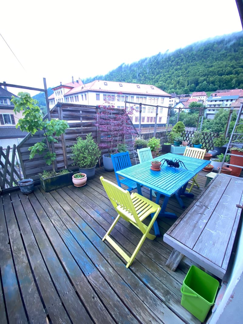 wooden deck, blue table, yellow chairs, potted plants, wooden bench, green bucket, distant buildings, mountain