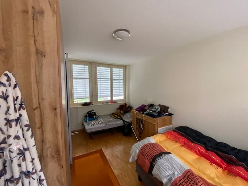 Bedroom with wooden floor, white walls, bed, small bed, window with blinds, wooden cabinet, rug, ceiling light