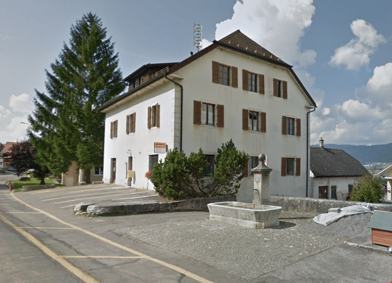 white building with brown windows and shutters, cobblestone driveway, parking area, small bush in front, and a fountain