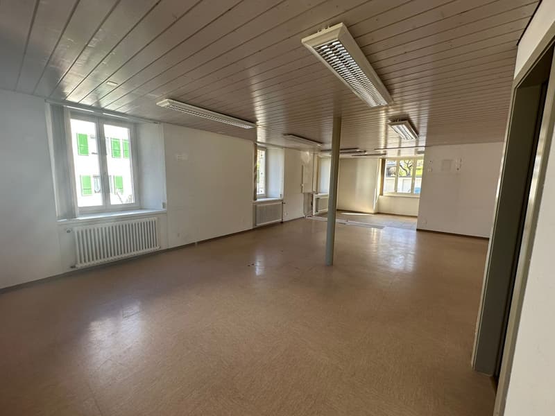 Empty room with white walls, wooden ceiling, windows, radiators, and a central pole.