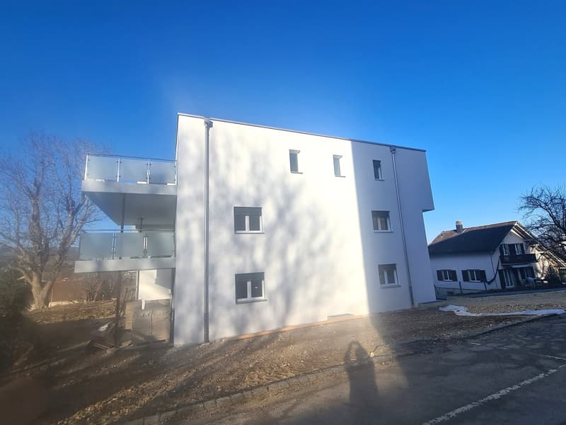 Newly constructed white house, 3 stories, windows with white frames, balconies, located in a neighborhood with other houses, partly unfinished area in front of the building.