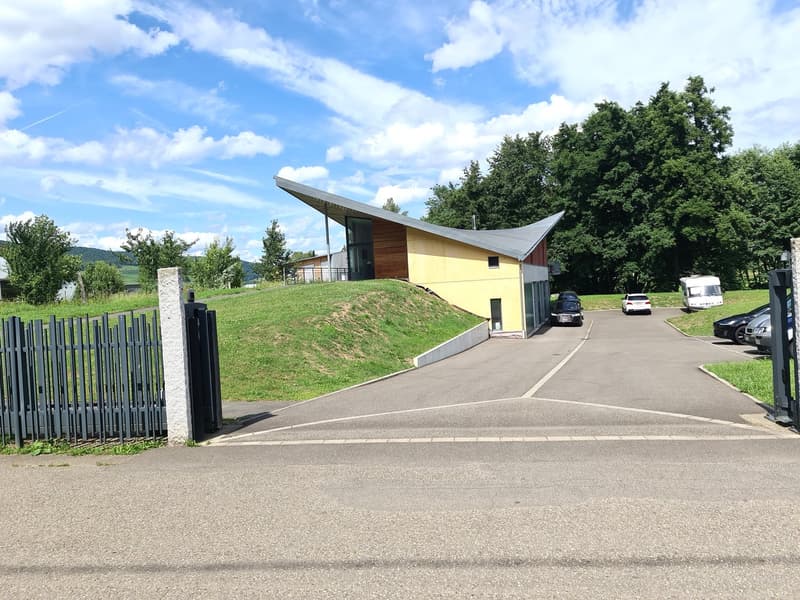 modern single-story building, angled roof, yellow exterior, wooden accents, raised ground floor, fenced entrance, parked cars, sloped driveway