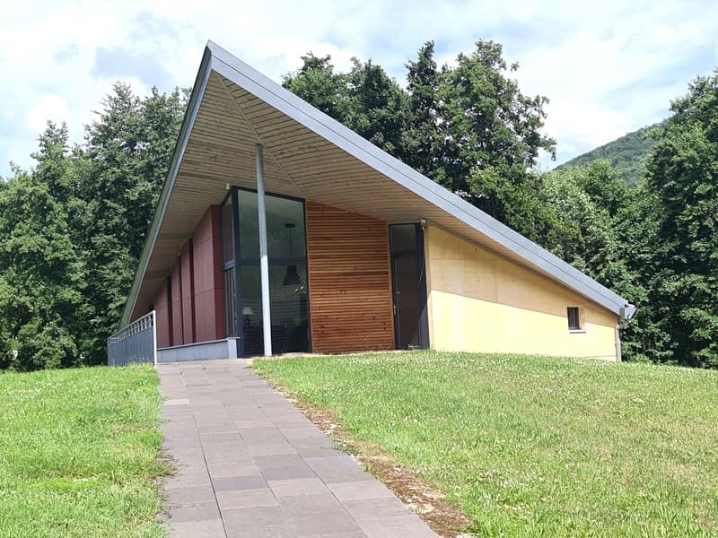 Architectural building with triangular roof, wooden walls, glass windows, elevated ground, green surroundings