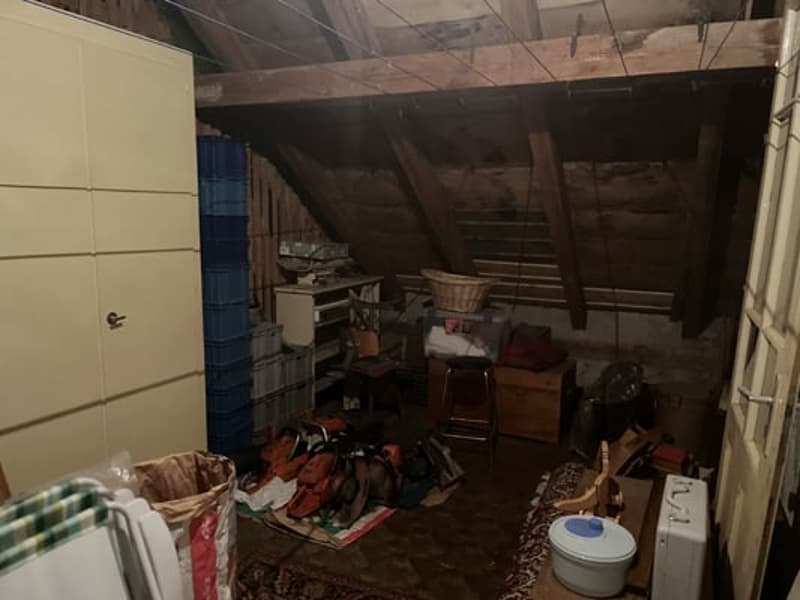 Cluttered attic space with various items and storage boxes, exposed wooden beams and rafters in the ceiling.