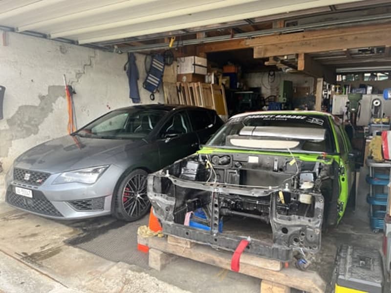 The image shows a garage with two cars undergoing repairs or maintenance. One car appears to be a grey Seat Leon, while the other is a green vehicle that is partially disassembled, likely for repairs or modifications.