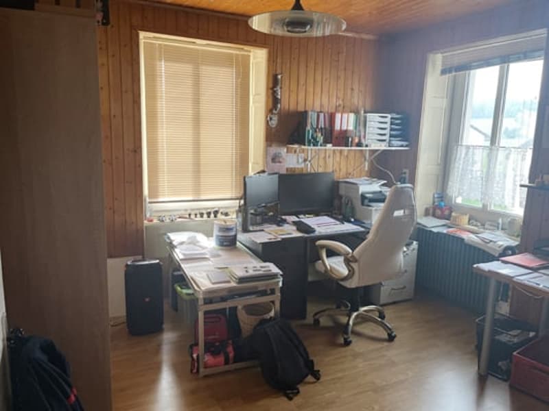 The image shows a cluttered home office space with a desk, computer, shelves, and various office supplies and equipment. The room has wooden paneled walls and a window providing natural light.