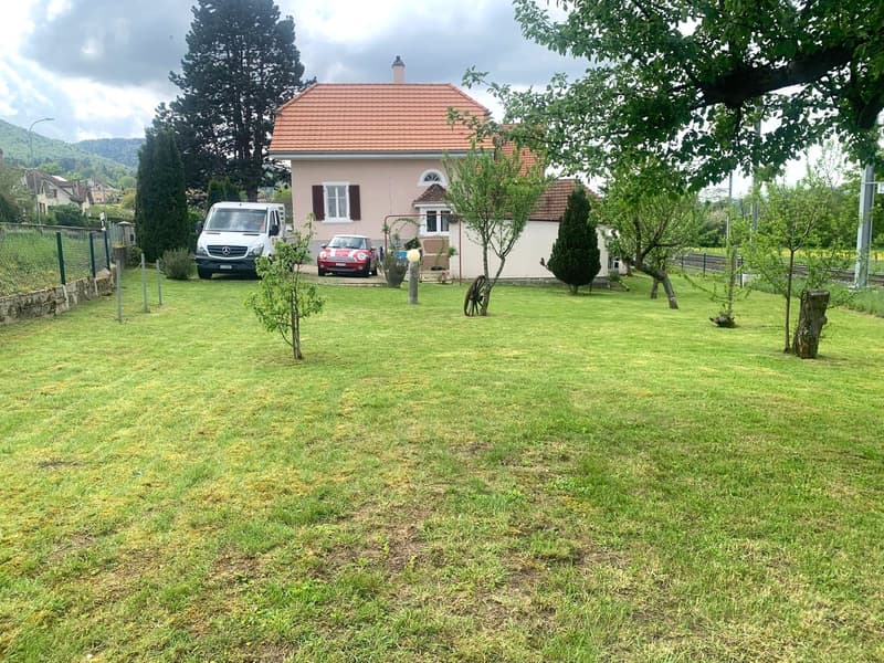 2 story house, pink exterior, tiled roof, fenced yard, 2 cars parked, trees, and shrubs