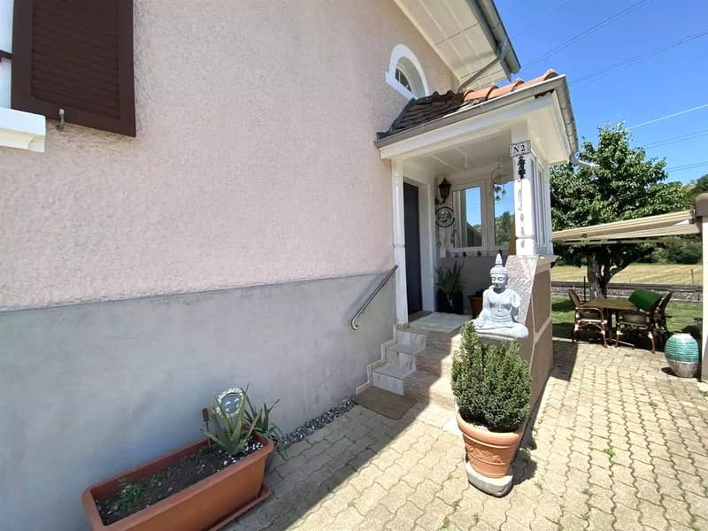 Exterior of a house with stone stairs, tiled walkway, and outdoor seating, a window, a door, and two potted plants. An awning on the right with a table and chairs, and a small tree behind it. A tree and bushes in the background.
