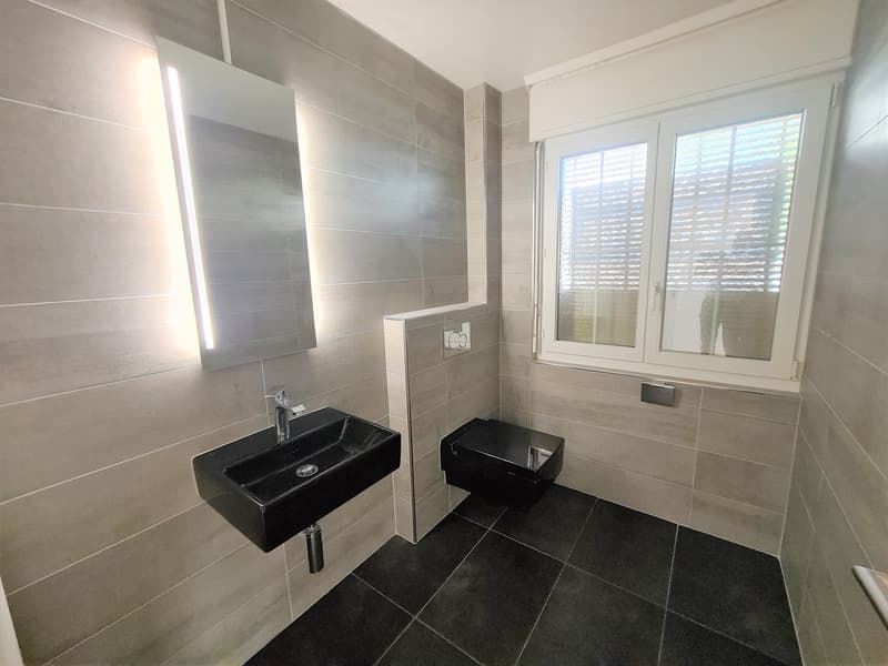 Spacious bathroom with gray tile walls, black tile floor, two black vessel sinks, and a window with shutters.