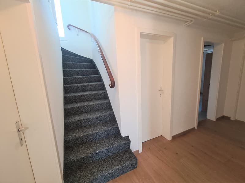 The image shows the interior of a building, with a staircase leading up to the second floor. The stairs are covered in a gray carpet, and there is a wooden handrail on the wall. The hallway on the ground floor has white walls and a wooden floor.