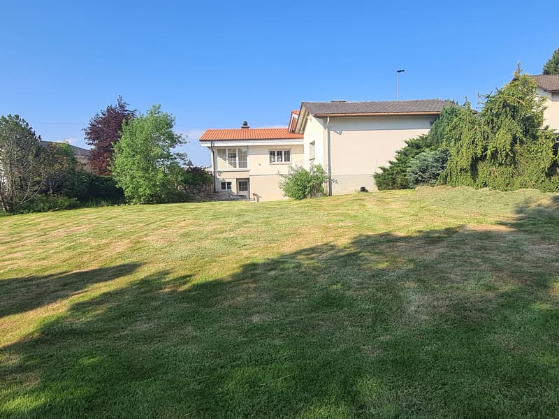 2-story house with a beige exterior, surrounded by a well-maintained lawn and landscaping, including trees and shrubs. The house has a tiled roof and appears to be in a residential neighborhood.