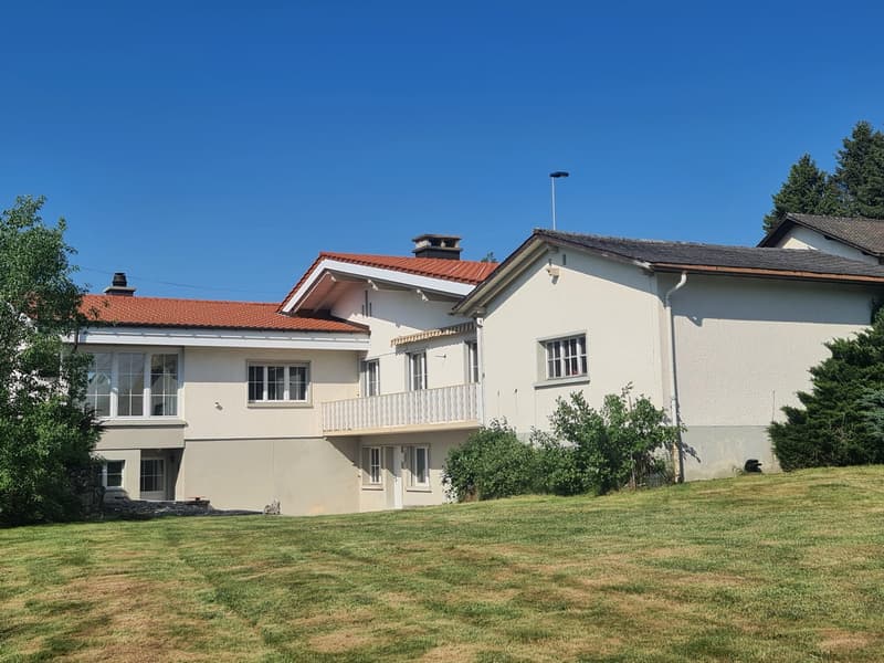 Two-story house with a red tile roof, white exterior walls, and large windows. The house has a balcony on the second floor and is surrounded by a grassy lawn and trees.