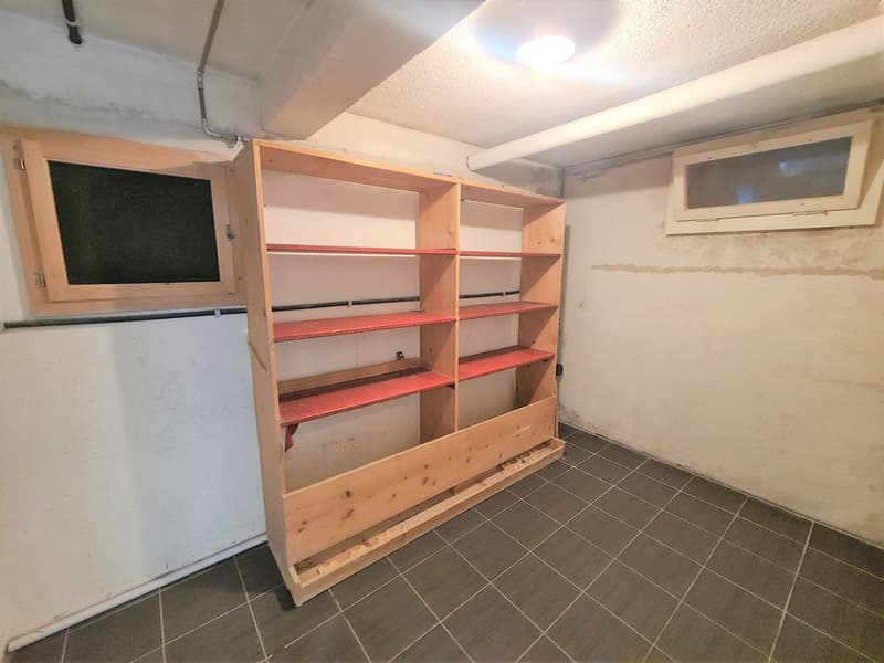 The image shows a storage room with a large wooden shelving unit. The room has a tiled floor and appears to be in a basement or garage area. The shelves are made of wood and have multiple levels for storage. There is also a small window and an air conditi