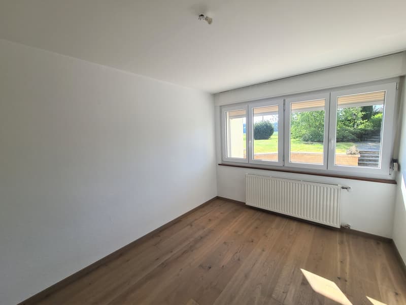 This image shows an empty room with white walls and a large window overlooking a garden. The room has a hardwood floor and a radiator along one wall.