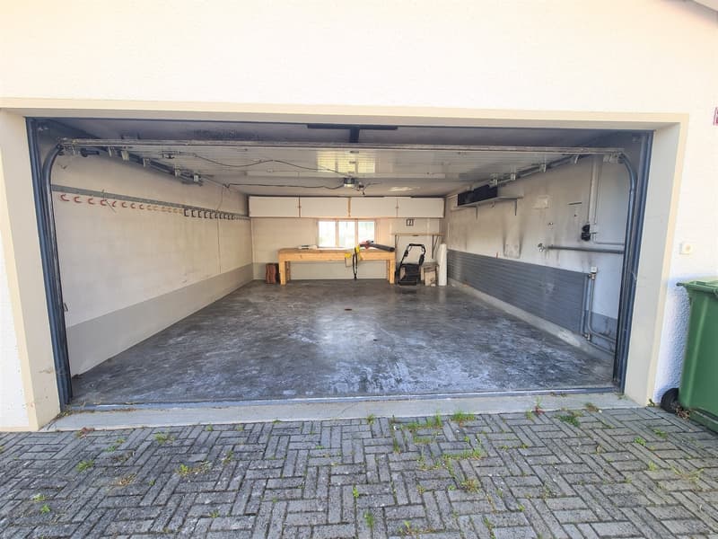 The image shows a large open garage space with a concrete floor. There are various tools and equipment visible inside the garage, including a workbench. The garage door is open, revealing the exterior brick paving outside.
