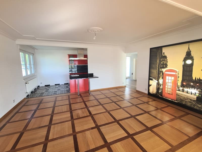 Open-concept living room with hardwood flooring in a checkered pattern, white walls, a red kitchen with built-in appliances including a dishwasher, and a large framed artwork depicting a London street scene.