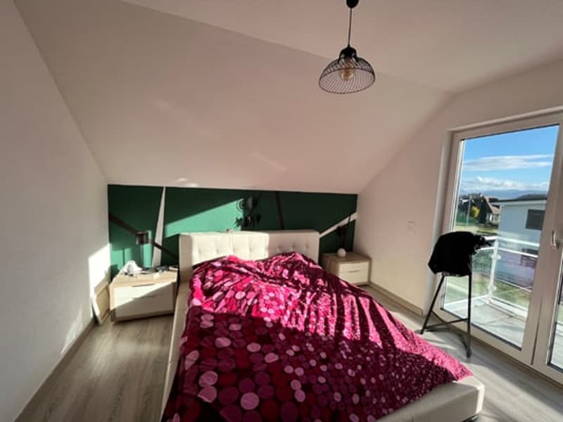 Spacious bedroom with large windows, hardwood flooring, and a pink and white patterned comforter on the bed. There is a balcony or terrace visible through the windows.