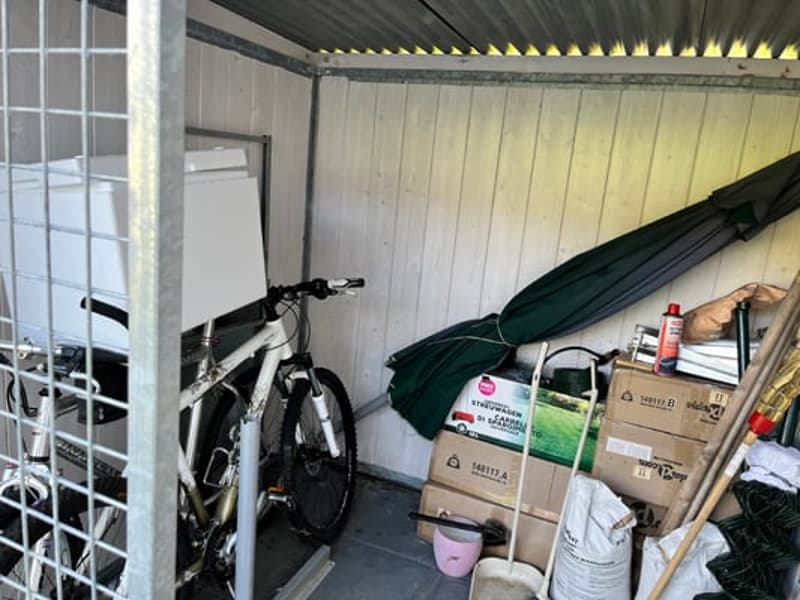 The image shows a cluttered garage space with various items stored, including a bicycle, boxes, and other miscellaneous items. The garage appears to have a corrugated metal roof and walls.