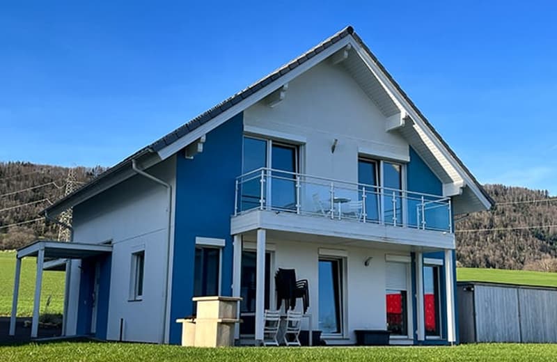 Two-story house with white exterior and blue accents, large balcony on the second floor, surrounded by a grassy yard