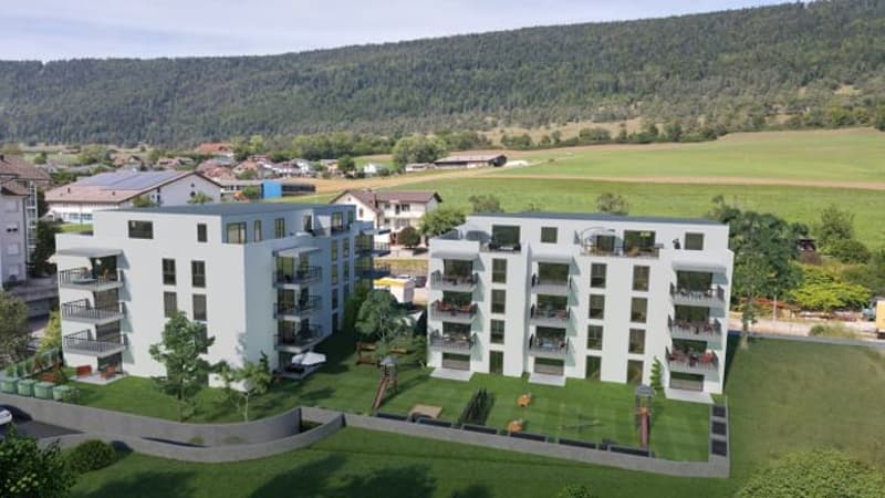 Two white apartment buildings, each with multiple floors and balconies, landscaped yard with play area, surrounding houses and greenery, scenic mountain view in the distance