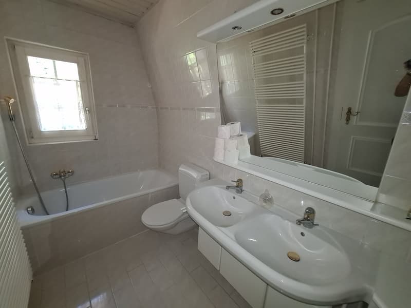 Spacious bathroom with white tiles, a bathtub, a sink with double vanity, and a towel rack. There is a window providing natural light and a private washing machine is visible.