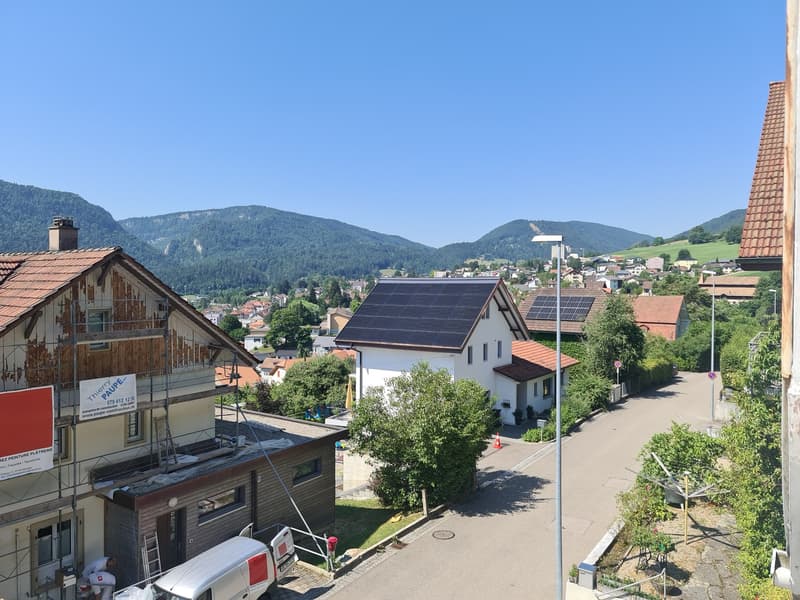 The image shows a residential area in a mountainous region. There are several houses with red tile roofs, some with solar panels installed. The houses are surrounded by trees and greenery, and there are paved roads and parking spaces visible. The backgrou