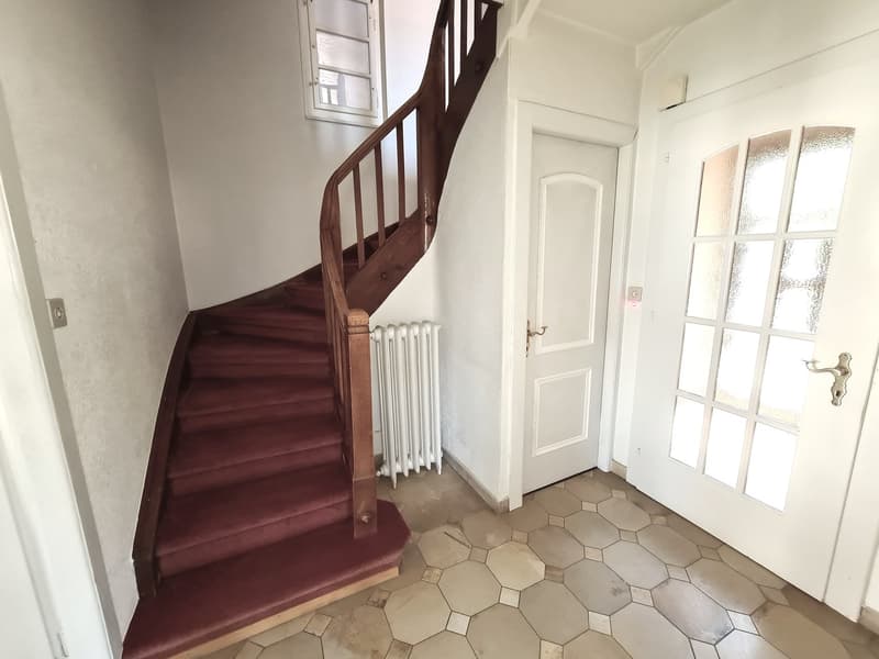 The image shows the entryway of a property, with a wooden staircase leading up to the second floor. The floor is covered in hexagonal tiles, and there is a white door leading to another room. The walls are painted a light color, and there is a window that