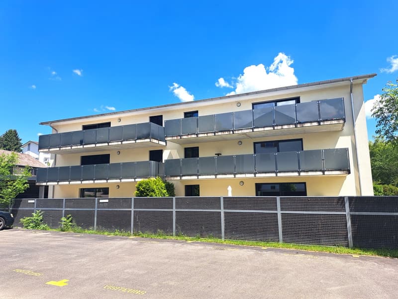 3-story apartment building with beige exterior, black balconies, and a fenced-in parking area in front.
