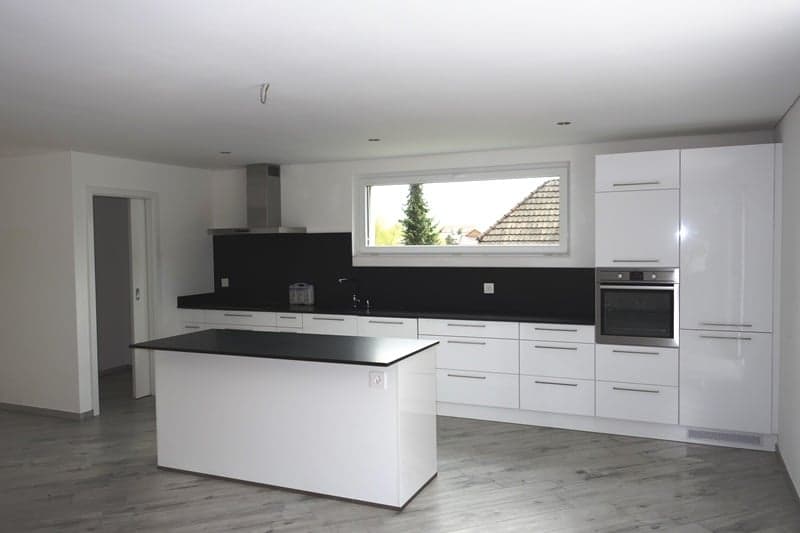 Spacious modern kitchen with white cabinets, black countertops, and stainless steel appliances including a dishwasher. Large windows provide natural light and a view outside.