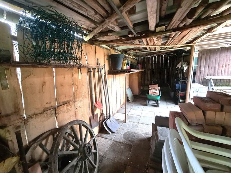 A wooden storage shed filled with gardening tools, a wheelbarrow, stacked bricks, and a bucket.