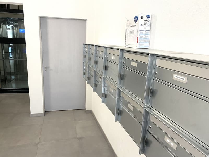 Mailboxes mounted on a white wall, a door to the right, a glass door with black frame behind, polished floor
