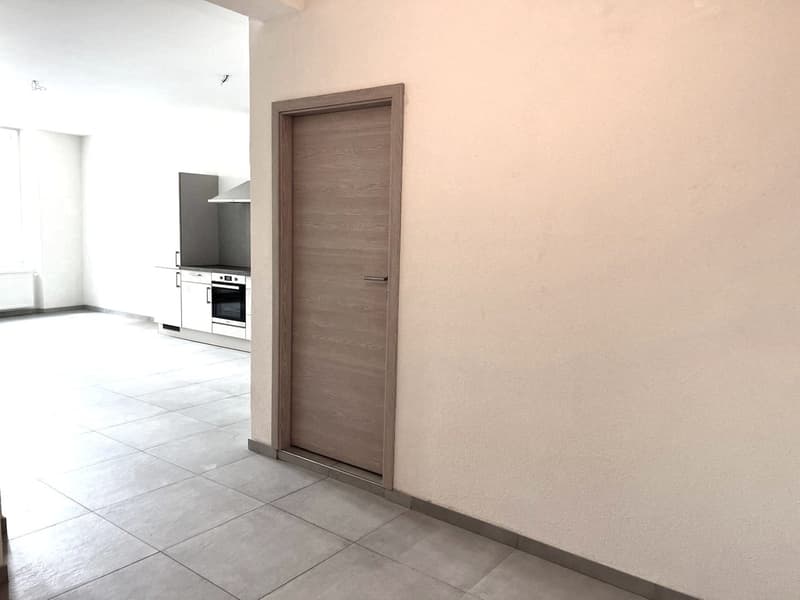 empty room with wooden door, tiled floor, adjacent to a kitchen with oven and cabinets