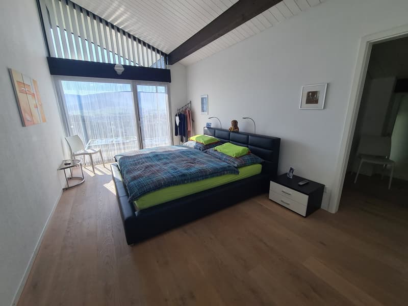 Large bedroom with a bed, bedside tables, picture frames, and a white chair in the corner. The room features wooden flooring, white walls, a sliding glass door leading to a balcony, and a large window overlooking the valley.