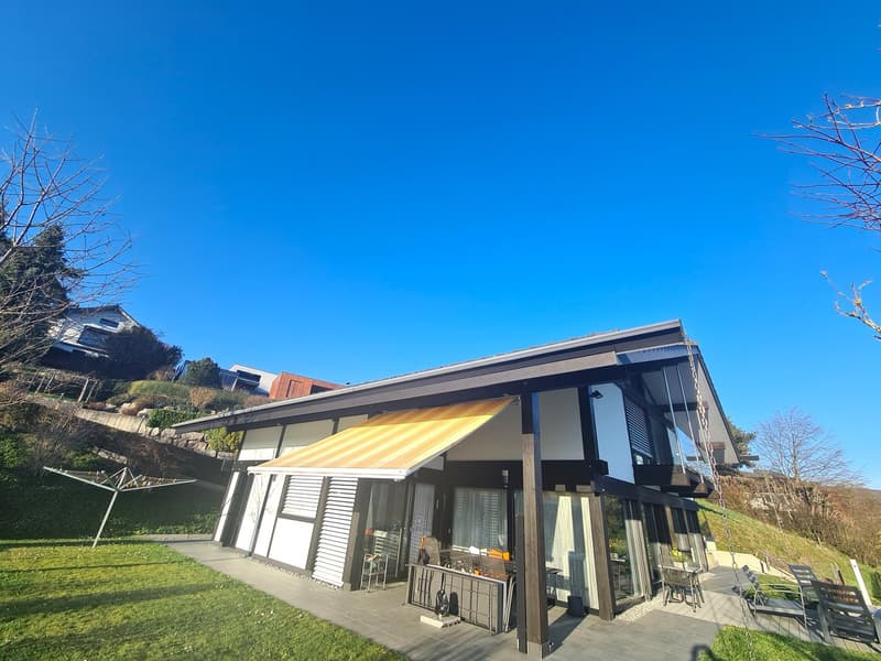 modern house with sloped roof, retractable awning, situated on a hill, paved patio, tables and chairs, distant view of trees and a house