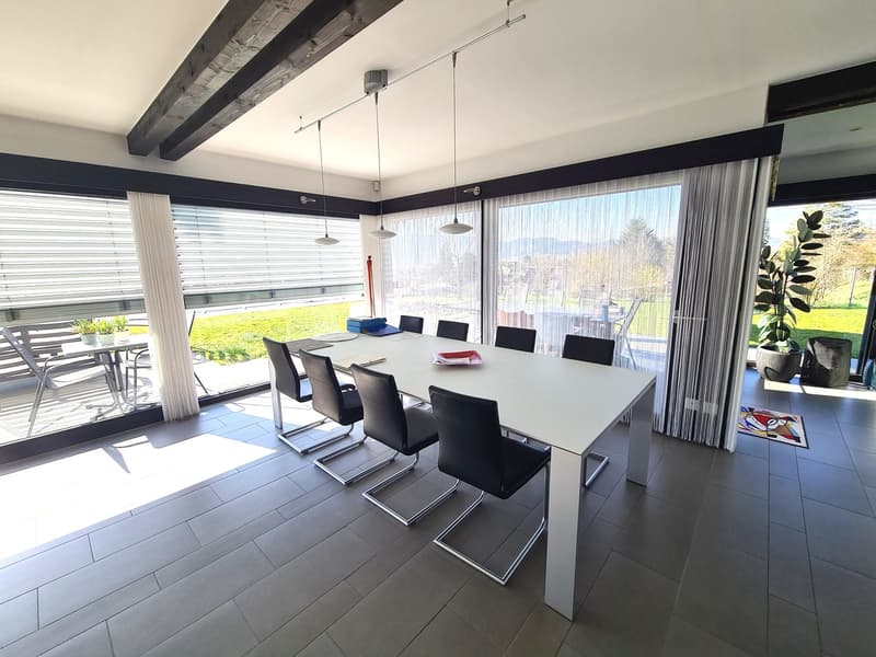 Modern dining room with glass doors, a white table with 8 chairs, a glass wall showing a balcony with chairs, a wooden ceiling, plants