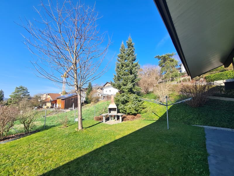 A well-kept garden featuring a grassy area with trees and a green lawn, a built barbecue pit in the center, and a view of a beautiful, clear blue sky