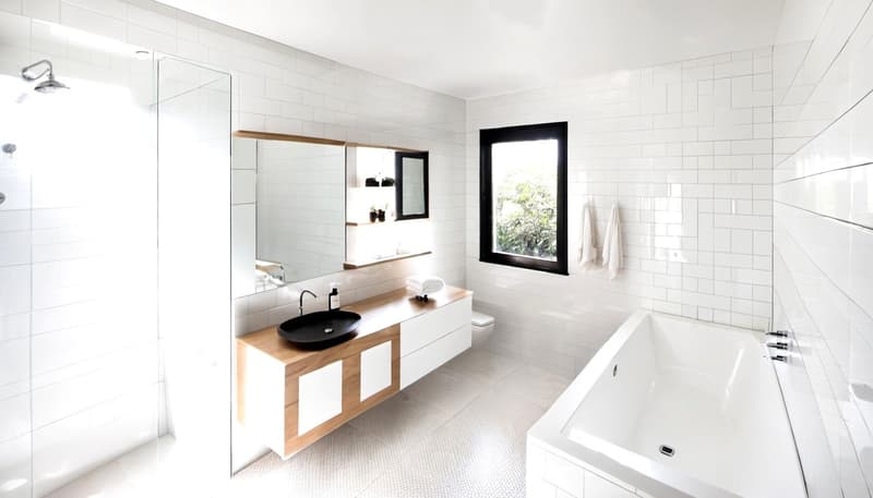 Modern bathroom with white tiles, large mirror, wooden vanity, large tub, shower enclosure, white toilet, shelves, faucets