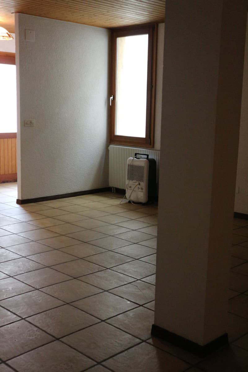 The image shows a room with white walls, a wooden ceiling, and a tiled floor. There is a window and a heating unit visible in the corner of the room.