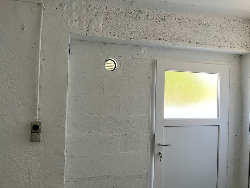 The image shows a simple, unfinished room with a white door and a small window. The walls are covered in a rough, gray plaster material. There is a ventilation duct or electrical outlet visible on the wall.