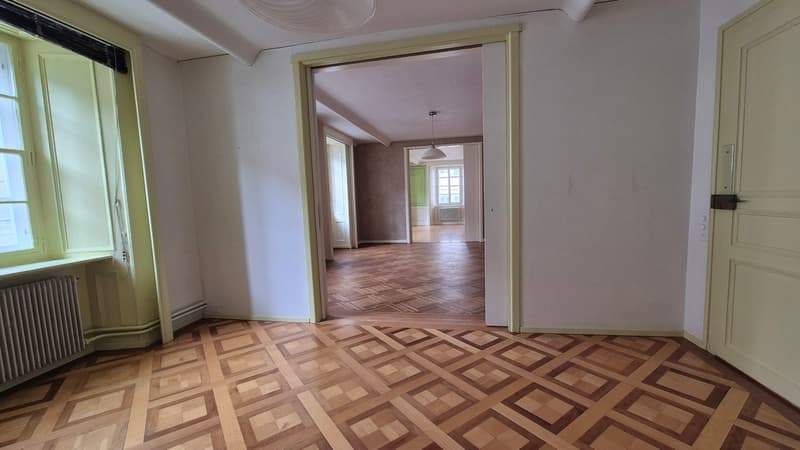 Checkered wooden floor, white walls, two doors, hallway leading to other rooms, natural light from windows, radiators, ceiling light fixture