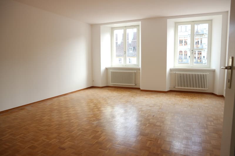 Empty room with wooden parquet flooring, white walls, two arched windows, and white radiators.