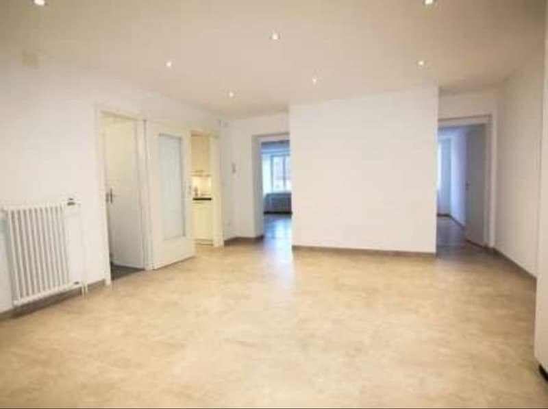 Empty room with light beige walls and floor, a white radiator and three doors leading to different rooms.