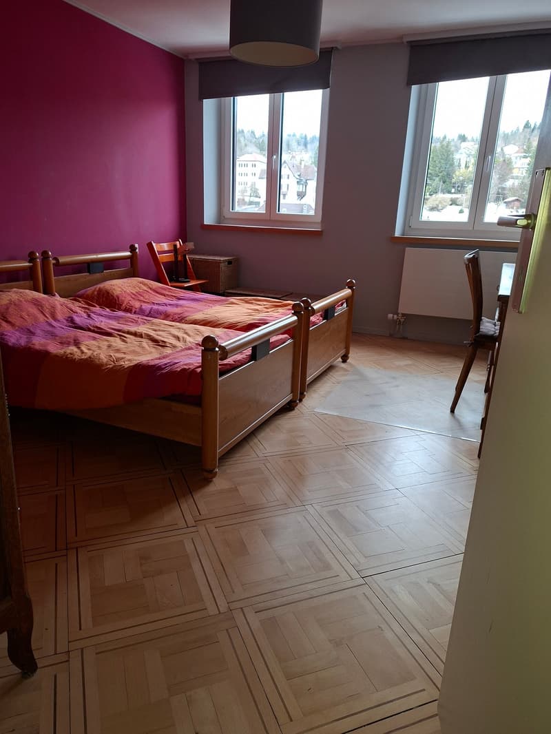 two beds, pink wall, wooden floor, window, wooden chairs, radiator, rug